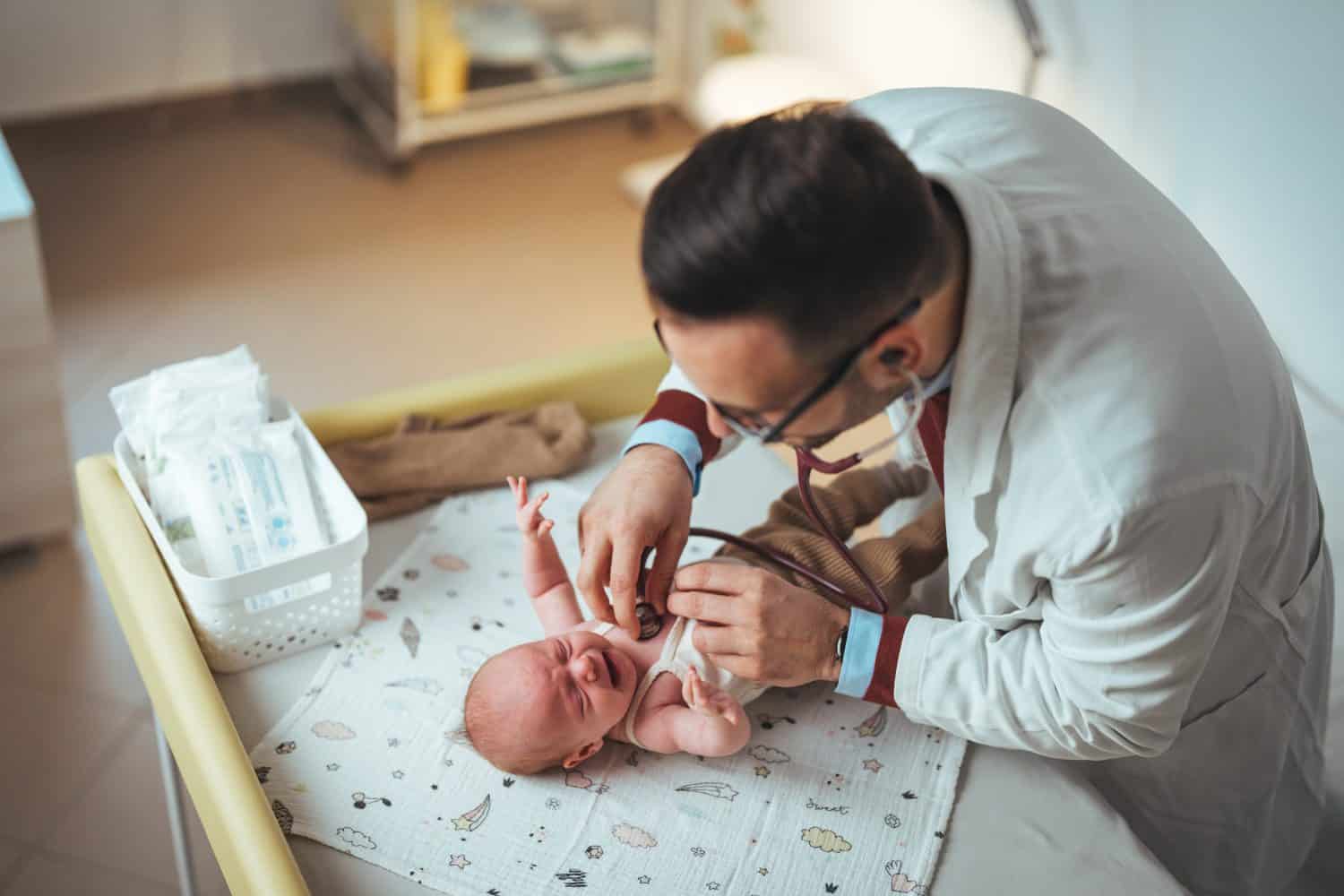 A professional pediatrician in medical attire gently examines a newborn baby lying on a hospital changing table, using a stethoscope for health assessment.