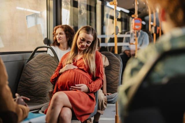 A pregnant woman in a red dress smiling and gently touching her belly while seated on a city bus during a commute.
