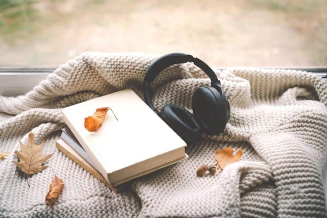 A pair of black headphones lies on an book, surrounded by autumn leaves and a knitted blanket, creating a warm and inviting atmosphere perfect for relaxation. Read, Rest. Cozy autumn or winter concept