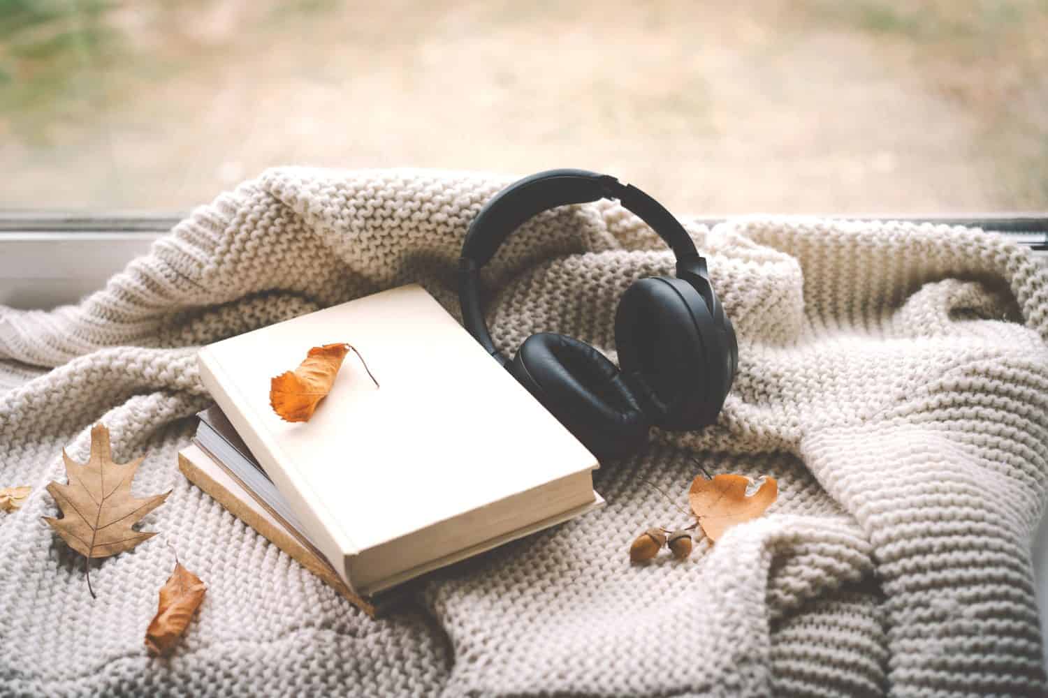 A pair of black headphones lies on an book, surrounded by autumn leaves and a knitted blanket, creating a warm and inviting atmosphere perfect for relaxation. Read, Rest. Cozy autumn or winter concept
