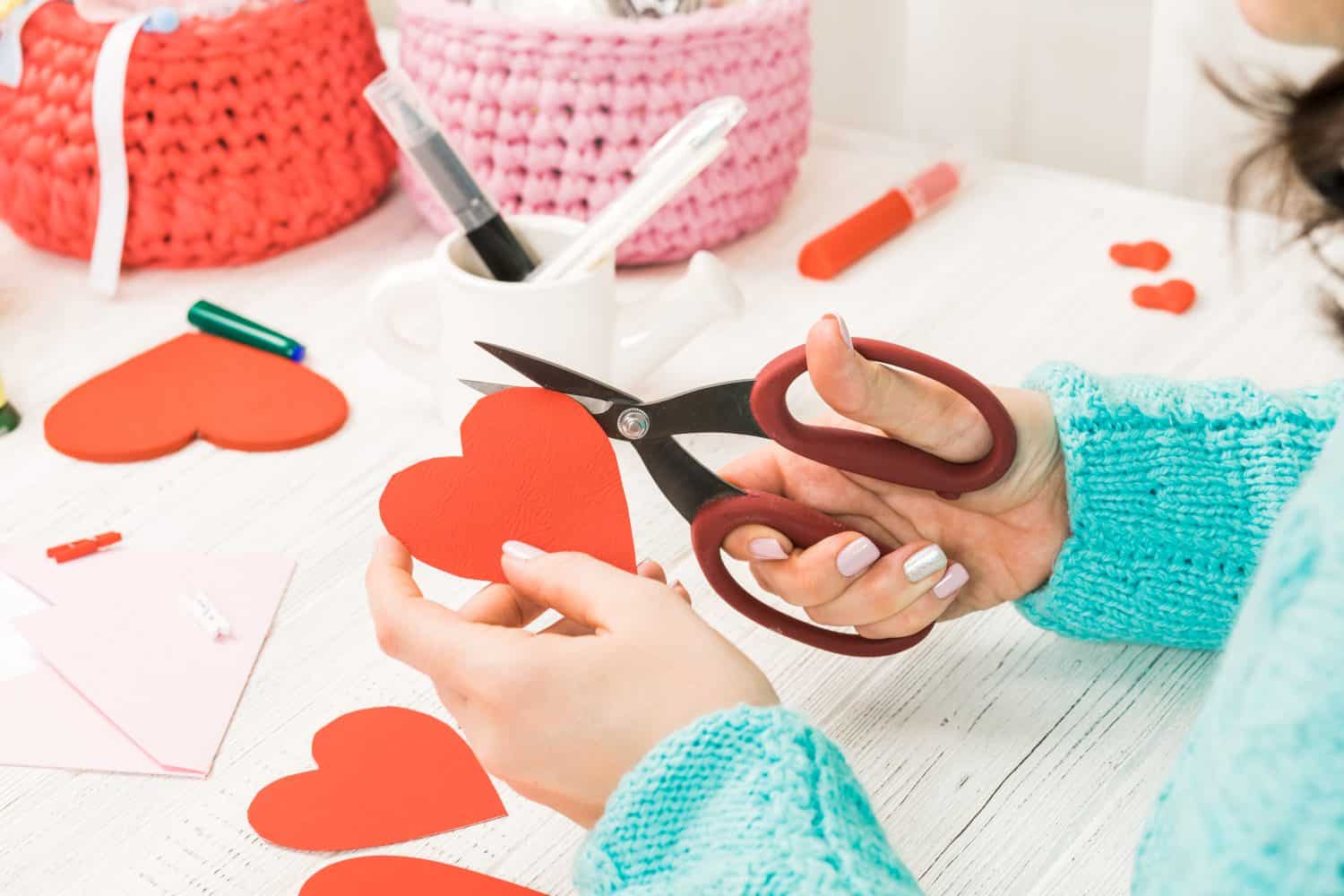 Valentine's day theme. Female hands cut the heart out of the paper. Packed gifts, tools on a battered wooden table. Workplace for the preparation of handmade ornaments