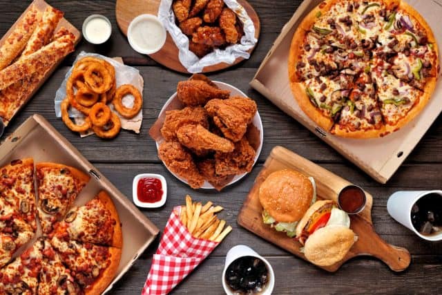 Buffet table scene of take out or delivery foods. Pizza, hamburgers, fried chicken and sides. Above view on a dark wood background.