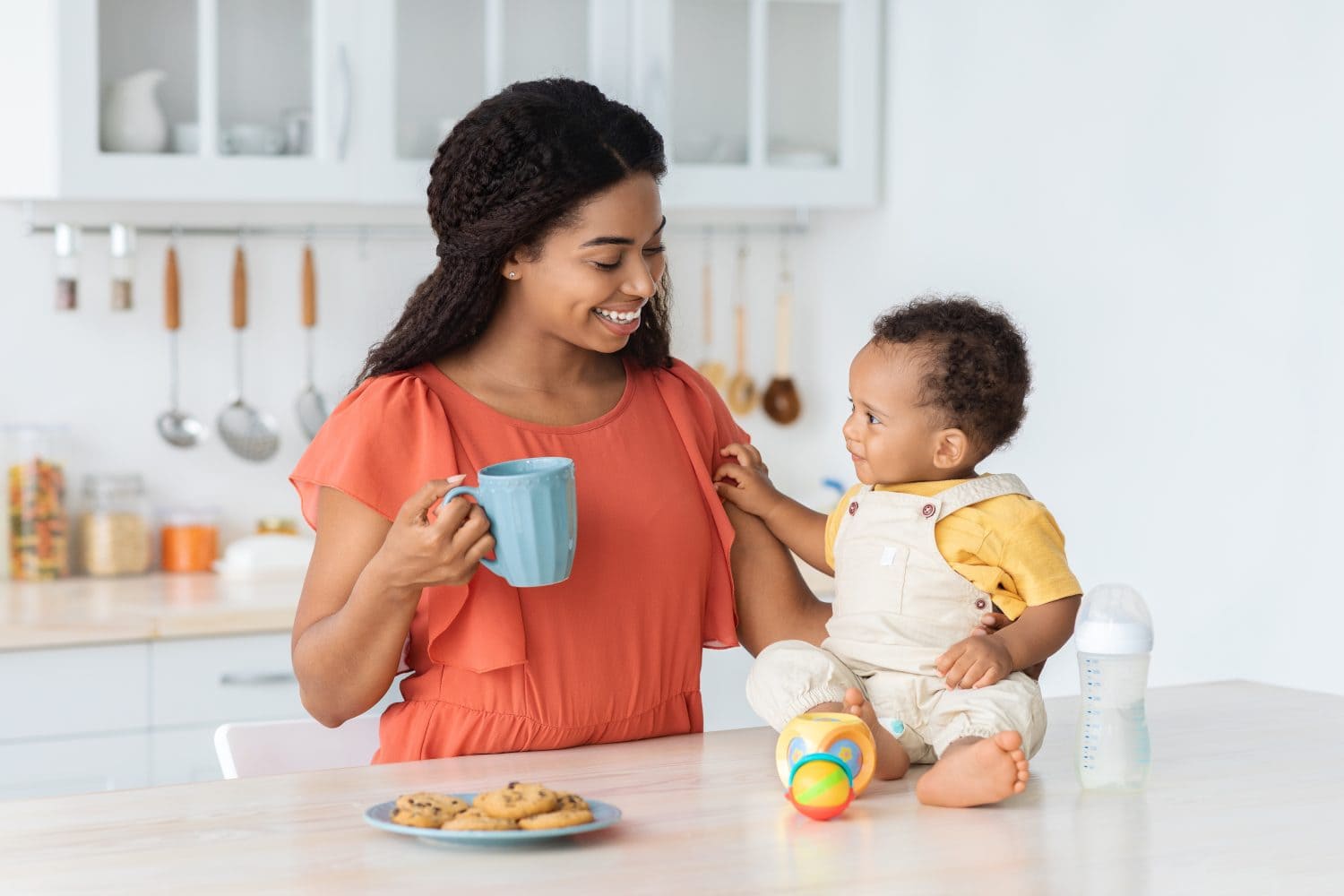 Time With Mom. Cute Little Black Baby Boy Relaxing With His Mother In Kitchen, Happy Young African American Mommy Drinking Coffee And Enjoying Playing With Her Adorable Infant Child At Home
