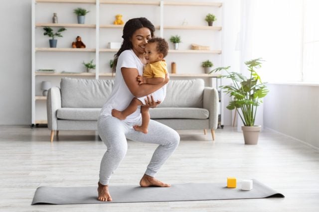 Postpartum Weight Loss. Happy Black Lady Making Squats With Baby On Hands While Training At Home, Smiling African American Woman Exercising In Living Room And Bonding With Infant Child, Copy Space
