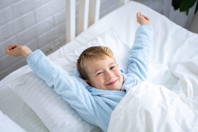 a cute boy in a blue bathrobe wakes up at home in the morning in a bed with white linens, rubs his eyes, yawns, smiles, stretches after sleep, a healthy sleep for children