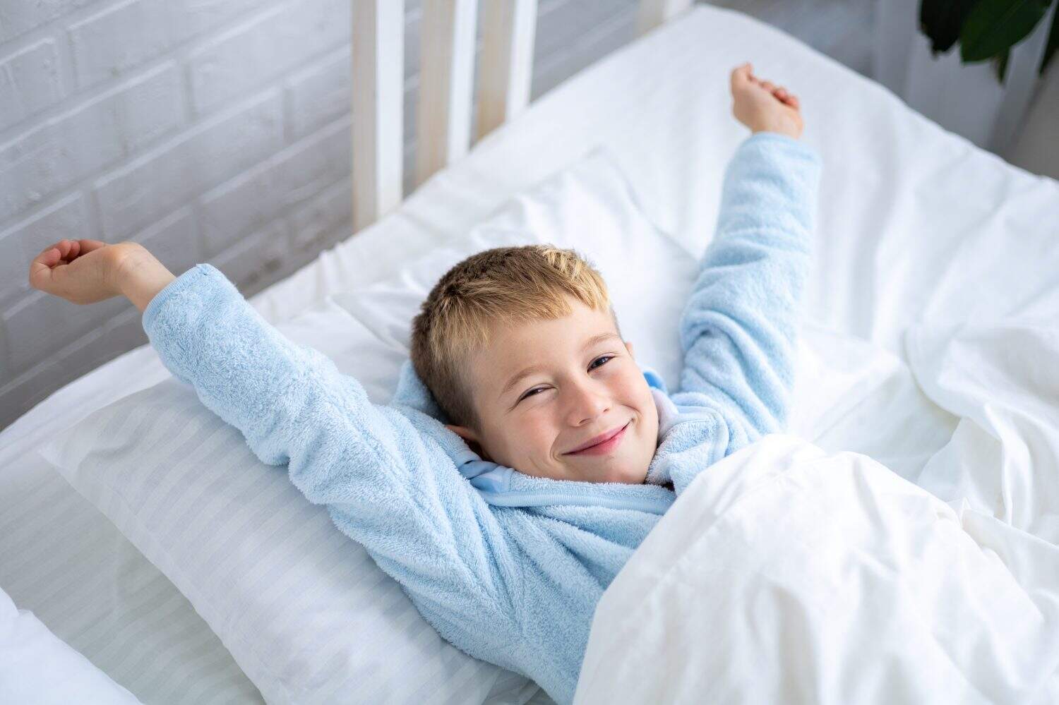 a cute boy in a blue bathrobe wakes up at home in the morning in a bed with white linens, rubs his eyes, yawns, smiles, stretches after sleep, a healthy sleep for children