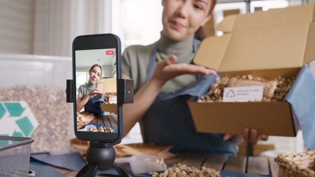 SME owner go green net zero retail store live stream talk on phone showing eco care plastic free packaging box Asia people young woman record video on reel shop happy side hustle