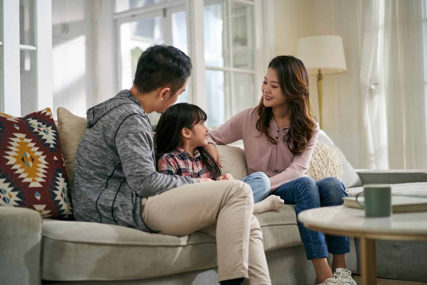 young asian mother and father sitting on family couch at home having a pleasant conversation with daughter