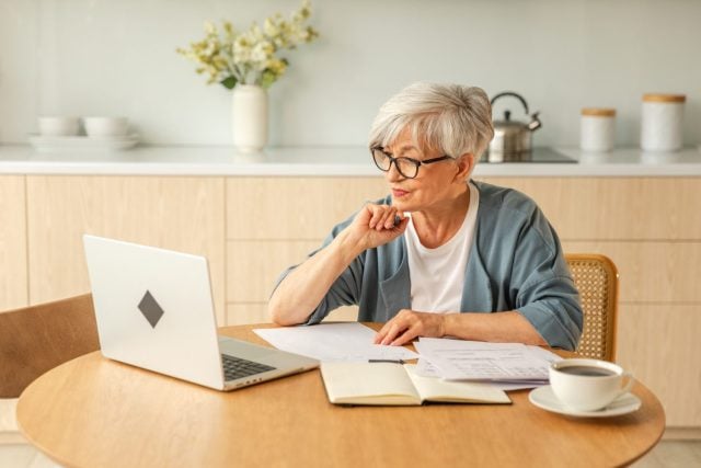 Online education courses webinar concept. Middle aged senior woman using laptop computer writing notes. Focused mature old woman enjoying studying online from home written records doing online work