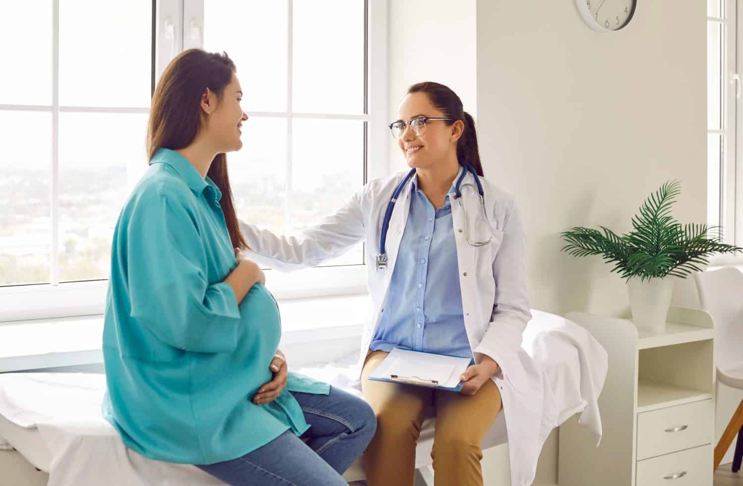 Friendly doctor supports pregnant woman patient at clinic. Smiling nurse or obstetrician with clipboard puts her hand on shoulder of young pregnant lady. Pregnancy, health care concept