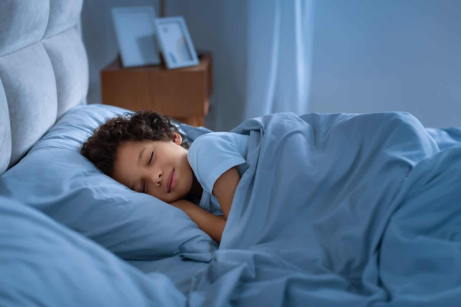 African American young boy peacefully sleeps in a bed with blue sheets, his eyes closed in a state of relaxation. The room is dimly lit with a warm glow, creating a cozy atmosphere