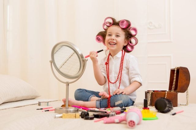 Happy child. Little pretty girl makes a make-up on a bed in the bedroom . Girl laughing with curlers on her head , and brushes for makeup in the hands