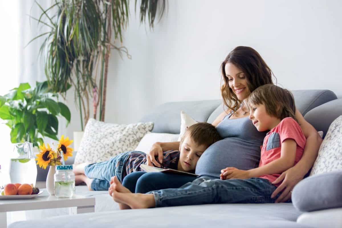 Young pregnant woman, reading a book at home to her two boys, eating fruits, hugging and laughing