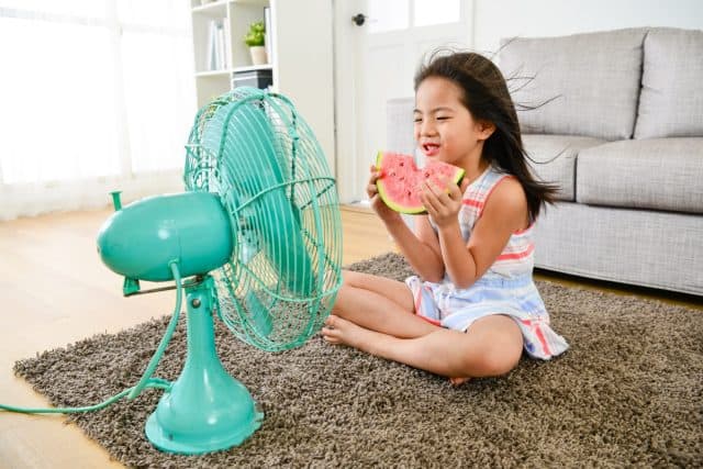 attractive young female little children sitting in front of electric fan blowing cooling wind and eating cold watermelon feeling refreshing during summer season.