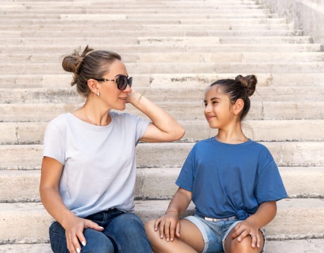 Middle-aged Mother and her kid daughter sitting on stone stairs outdoor wearing t-shirts and looking each other. Loving family spending time together outdoors in summer, t-shirts mockup