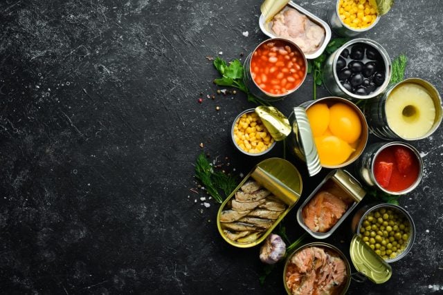 Canned vegetables, beans, fish and fruits in tin cans on black stone background. Food stocks.