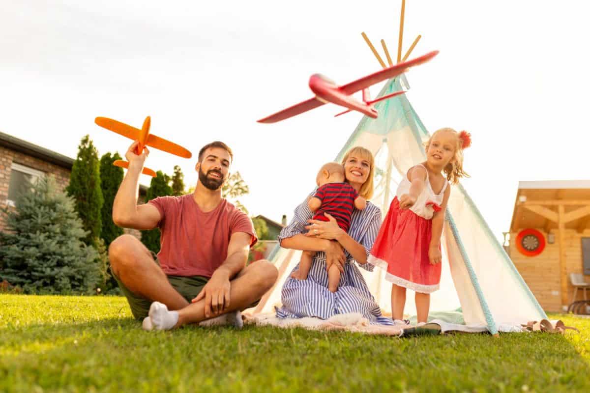 Beautiful young happy family having fun spending sunny summer day camping in the backyard, parents playing with their children, throwing toy airplanes