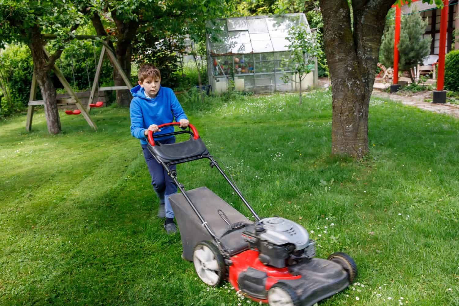 Happy preteen kid boy with lawn mower. Portrait of smiling teenager child working in garden, trimming grass. Garden works in summer. Kid helping