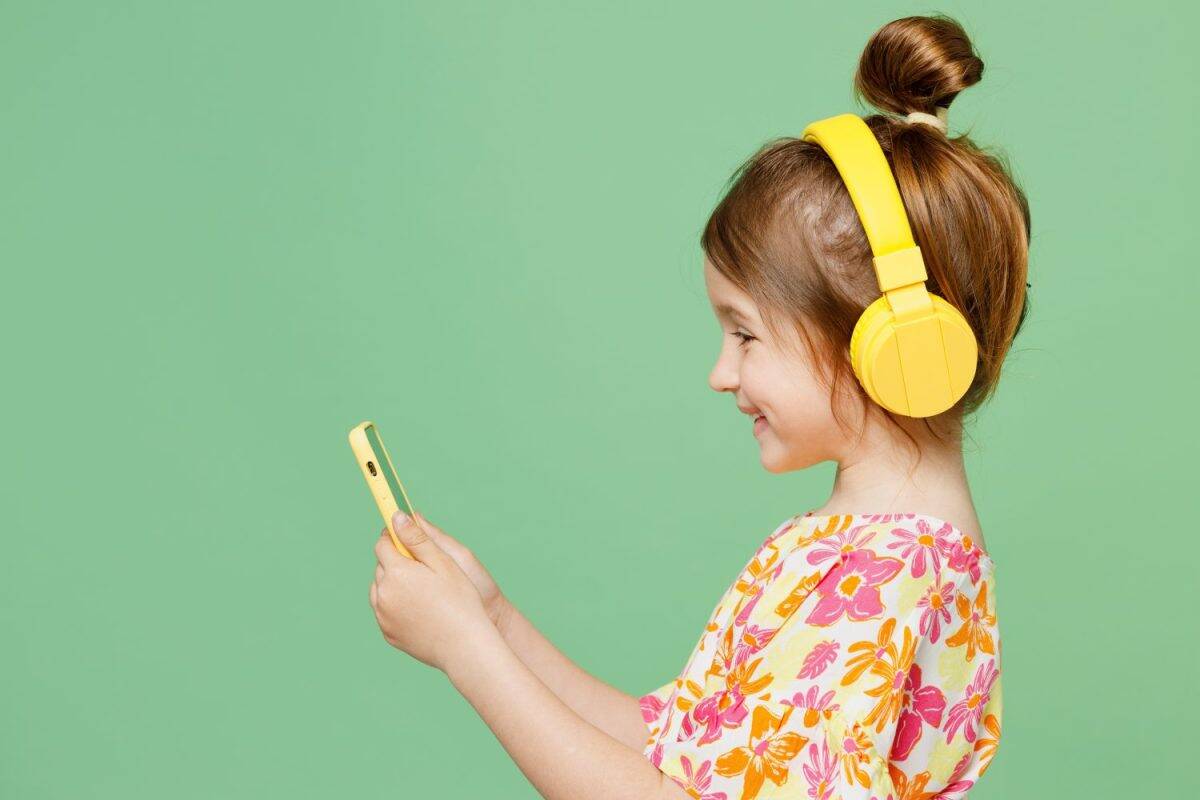 Side view little child kid girl 6-7 years old wear casual clothes headphones listen music use mobile cell phone isolated on plain pastel green background. Mother's Day love family lifestyle concept