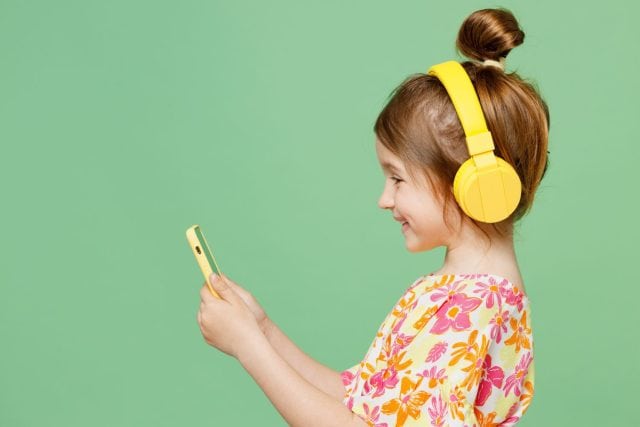Side view little child kid girl 6-7 years old wear casual clothes headphones listen music use mobile cell phone isolated on plain pastel green background. Mother's Day love family lifestyle concept