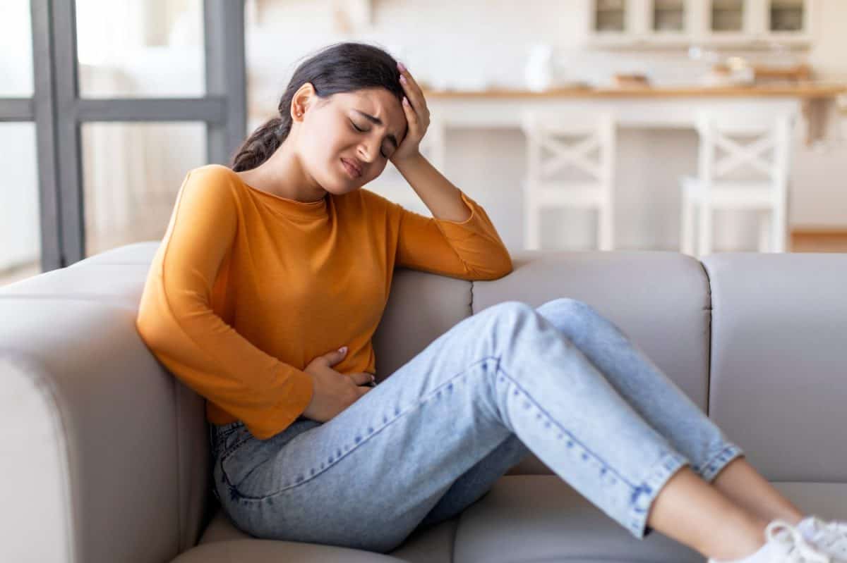 Sad young indian woman suffering from PMS and menstruation pain at home, upset eatsern female having stomach ache, acute abdominal pain and headache, sitting on couch in living room, copy space