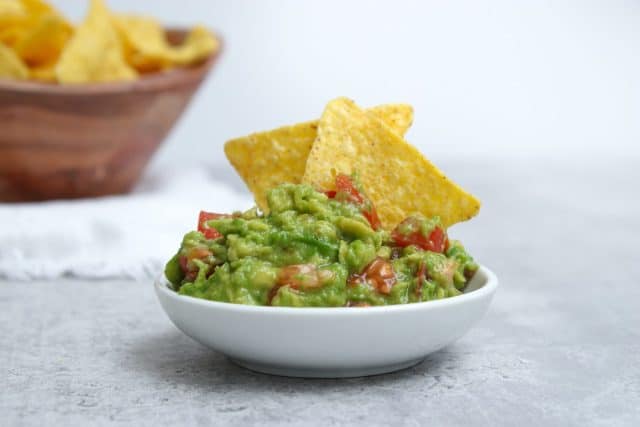 Close up of fresh avocado guacamole dip with salted tortilla chips in wooden bowl in background, grey marble table