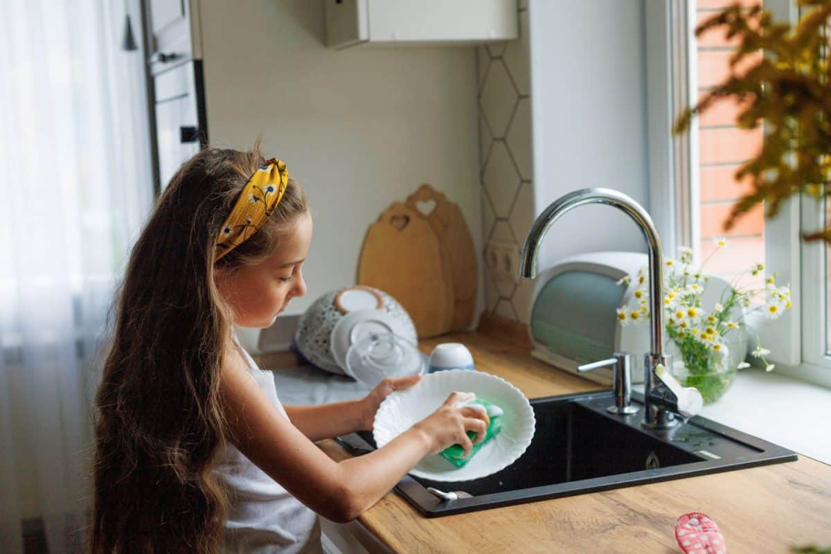 Cute little girl washing dishes with sponge in kitchen sink at home. Child doing chores concept.