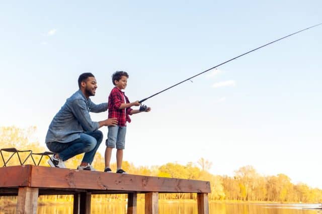 African American man and child sitting on a wooden pier holding fishing rods on the river, dad teaching his son to fish in the lake, family resting and relaxing on the weekend