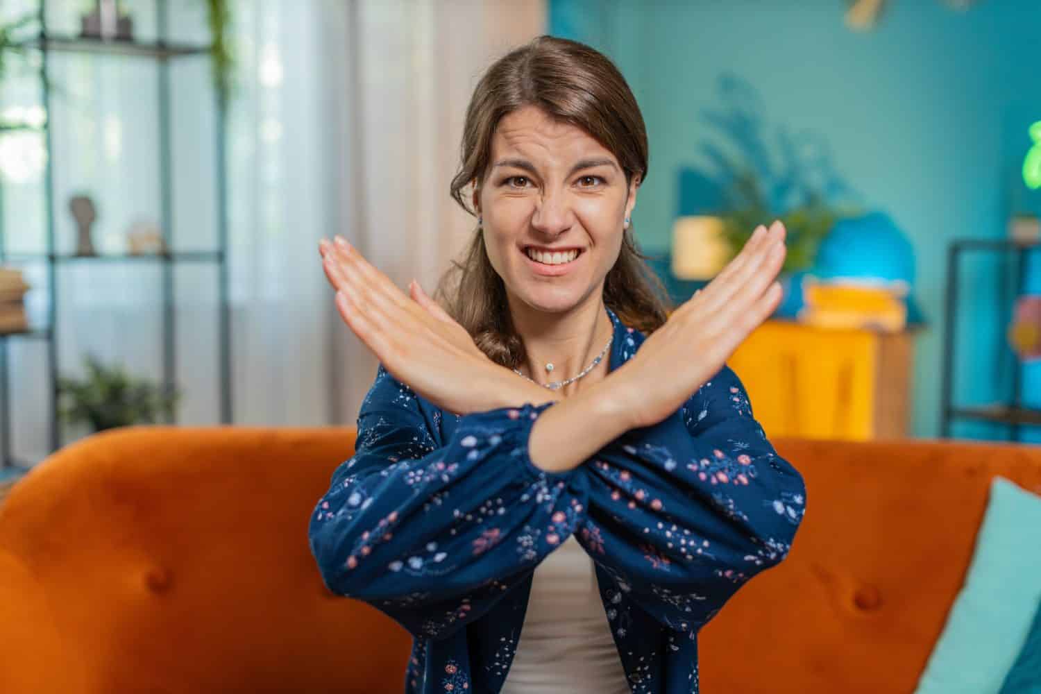 Stop gesture. Angry Caucasian woman says No hold palm folded crossed hands, warning of finish, prohibited access, declining communication, body language, danger. Girl at home room sits on sofa couch.