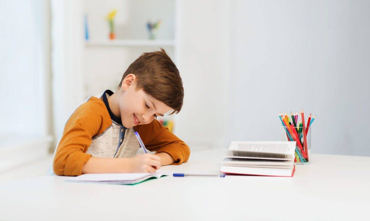 education, childhood, people, homework and school concept - smiling student boy with book writing to notebook at home