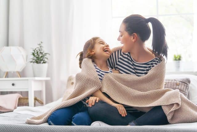A nice girl and her mother enjoy sunny morning. Good time at home. Child wakes up from sleep. Family playing under blanket on the bed in the bedroom.