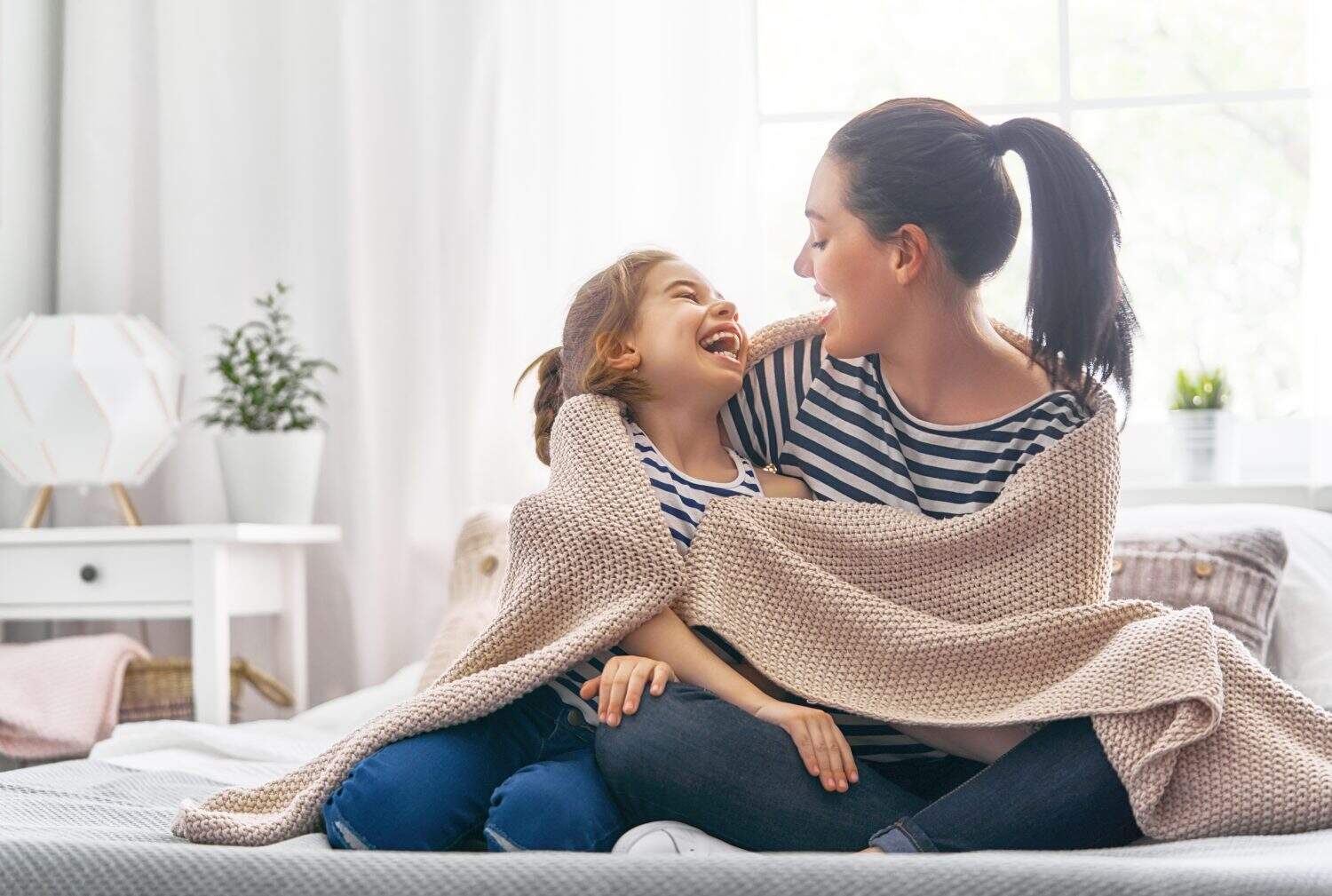 A nice girl and her mother enjoy sunny morning. Good time at home. Child wakes up from sleep. Family playing under blanket on the bed in the bedroom.