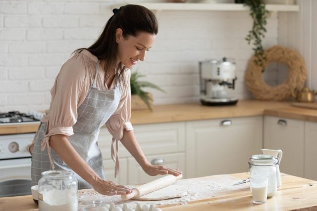 Happy attractive young adult woman lady housewife baker wear apron holding pin rolling dough on kitchen table baking pastry concept cooking cake biscuit doing bakery making homemade pizza at home