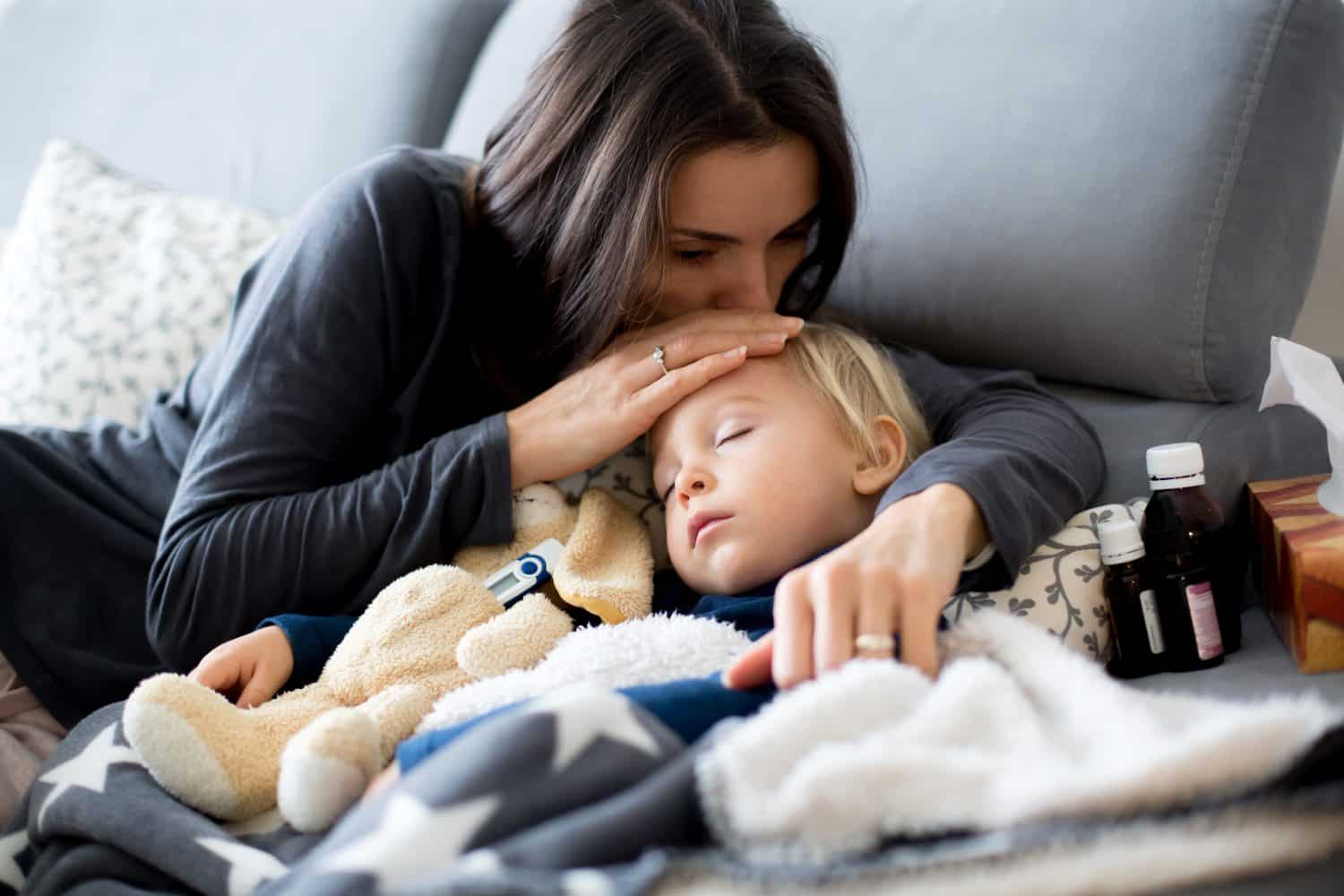 Blond toddler boy, sleeping on the couch in living room, lying down with fever, mom checking on him