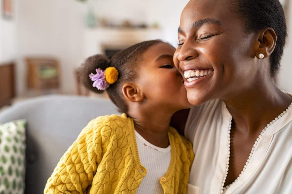 Close up of beautiful daughter kissing mother on cheek at home. African little girl giving kiss to happy mother. Lovely black female child kissing cheerful and proud woman on cheek for mother's day.