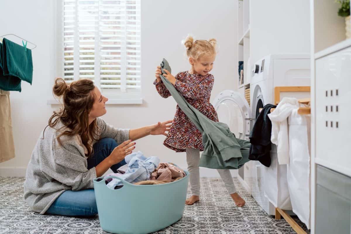 Mom spends time with daughter in bathroom, laundry room while doing daily chores, girl takes things out of washing machine and hands to woman who puts colorful clean clothes into bowl
