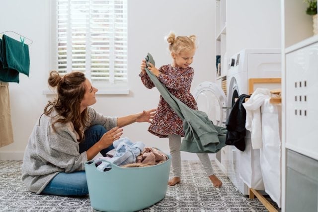 Mom spends time with daughter in bathroom, laundry room while doing daily chores, girl takes things out of washing machine and hands to woman who puts colorful clean clothes into bowl