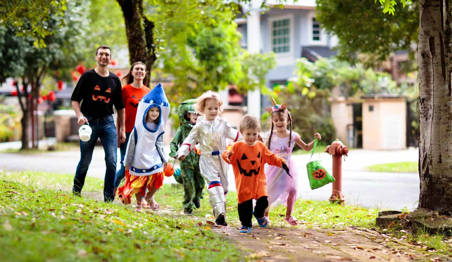 Child in Halloween costume. Asian and Caucasian kids and parents trick or treat on street. Little boy and girl with pumpkin lantern and candy bucket. Baby in witch hat. Autumn holiday fun.