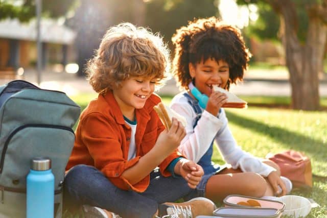 Charming schoolchildren after school resting sitting on the grass in the park. An African-American girl with a Caucasian boy took out lunchboxes with sandwiches from their backpack and eating.