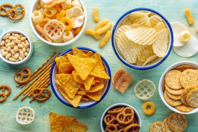 Salty snacks. Party food on a blue background. Potato and tortilla chips, crackers and other appetizers in bowls, overhead flat lay shot