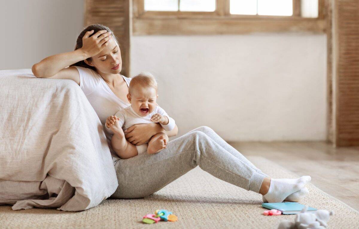 Tired infant baby cries in mother hands, depressed unhappy exhausted mom sitting on floor with crying little child on her lap, bedroom interior, copy space. Postnatal postpartum depression concept