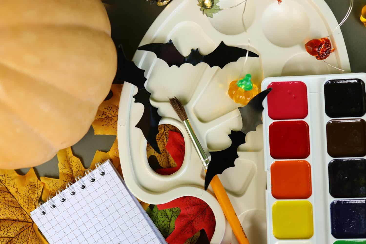 A table displays an assortment of Halloween crafting supplies including a painted pumpkin, bats, a palette with vibrant colors, and scattered leaves. Creativity fills the air.