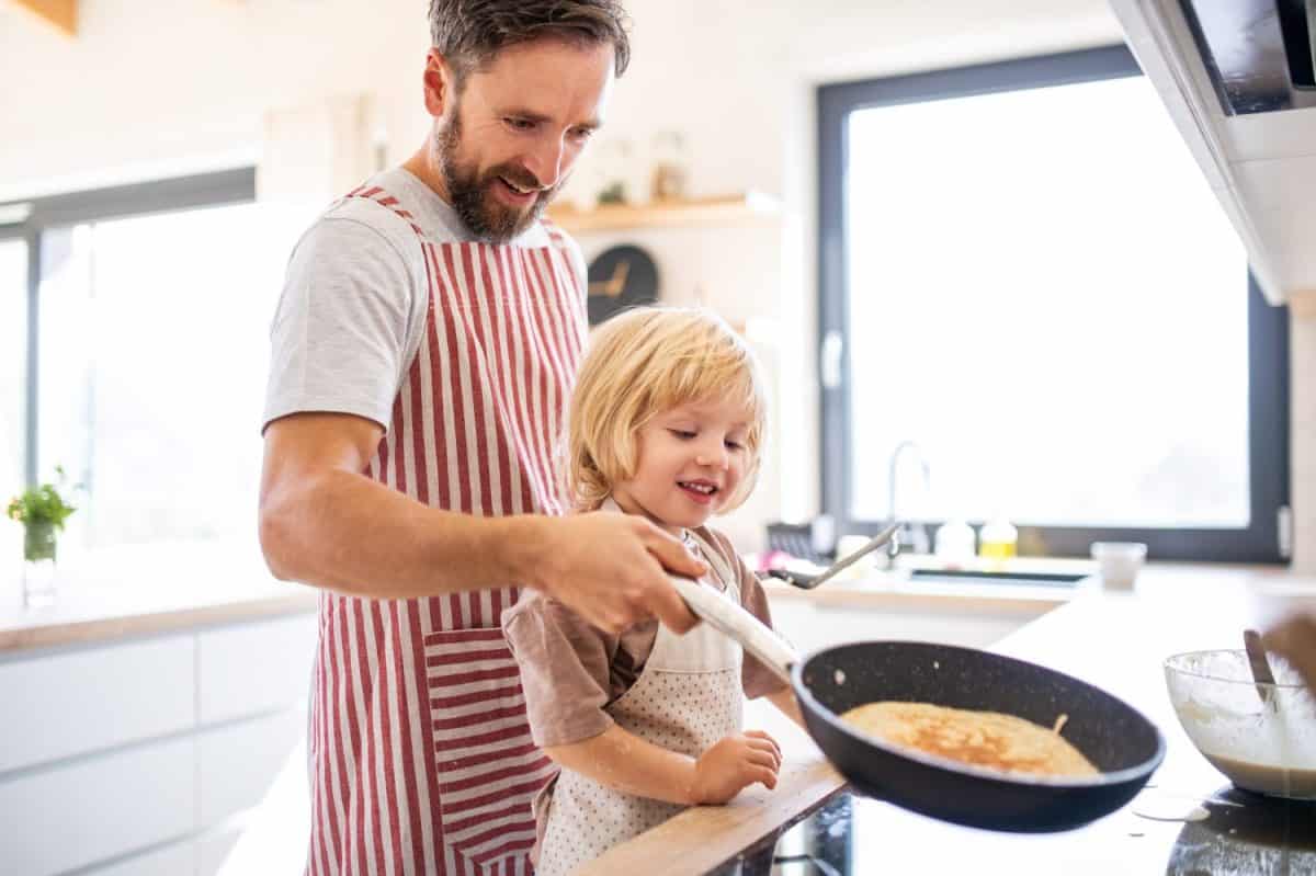 A side view of small boy with father indoors in kitchen making pancakes.