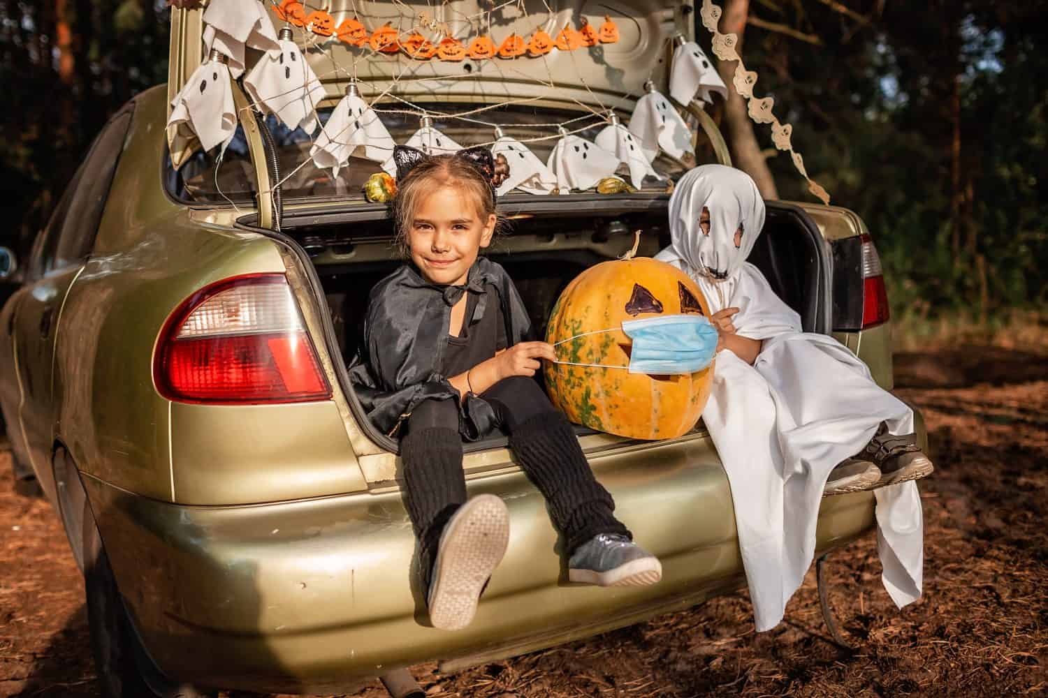 Alternative safe celebration. Cute kids celebrating Halloween party in the trunk of car with spider net, ghosts, carved pumpkin in medical mask for fun and other decoration, autumn outdoor