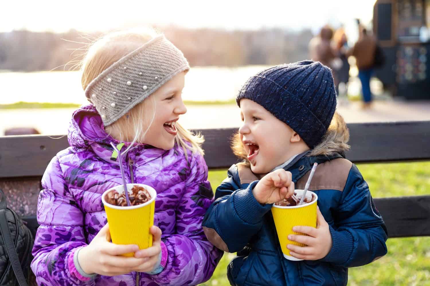 Two cute adorable siblings children sitting on bench drink delicious yummy hot chocolate, tea cocoa from paper cups during walk at city street park or backyard outdoors. Brother and sister enjoy fun