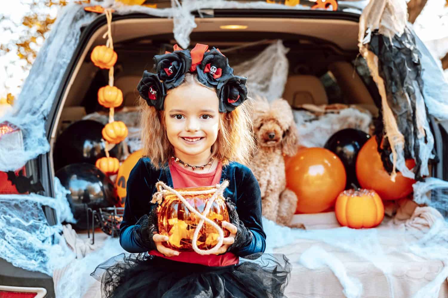 little girl in spooky costume and hat with bucket of sweets and cute poodle dog in ghost costume sits in trunk car decorated for Halloween with web, orange balloons and pumpkins, outdoor creative