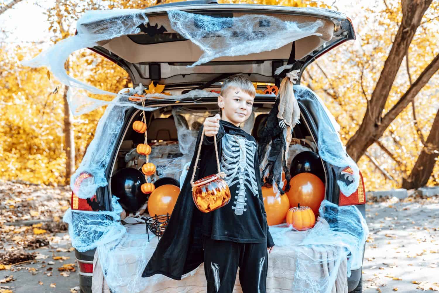 preteen cute caucasian boy in skeleton costume with bucket of sweets sits in trunk car decorated for Halloween with web, orange balloons and pumpkins, outdoor creative