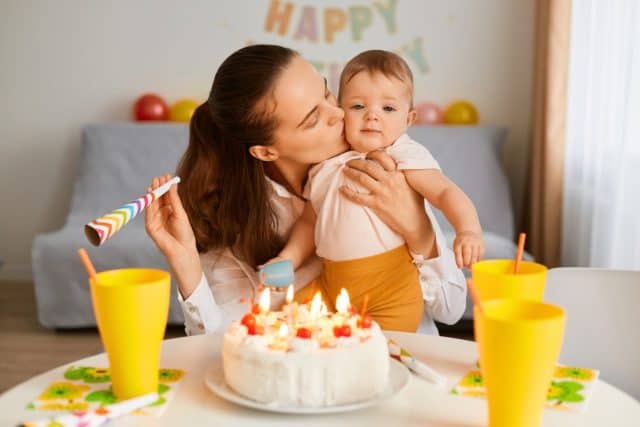 Indoor shot of dark haired woman with funny baby celebrating first birthday, sitting at table with birthday cake and drink, mother kissing her infant daughter, celebrating together.