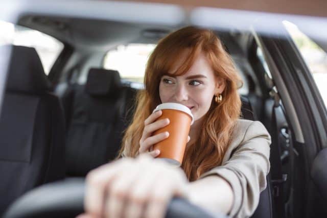 Happy young woman with coffee to go driving her car. Woman sipping a coffee while driving a car. Young woman drinking coffee while driving her car. Attractive red hair drives a car