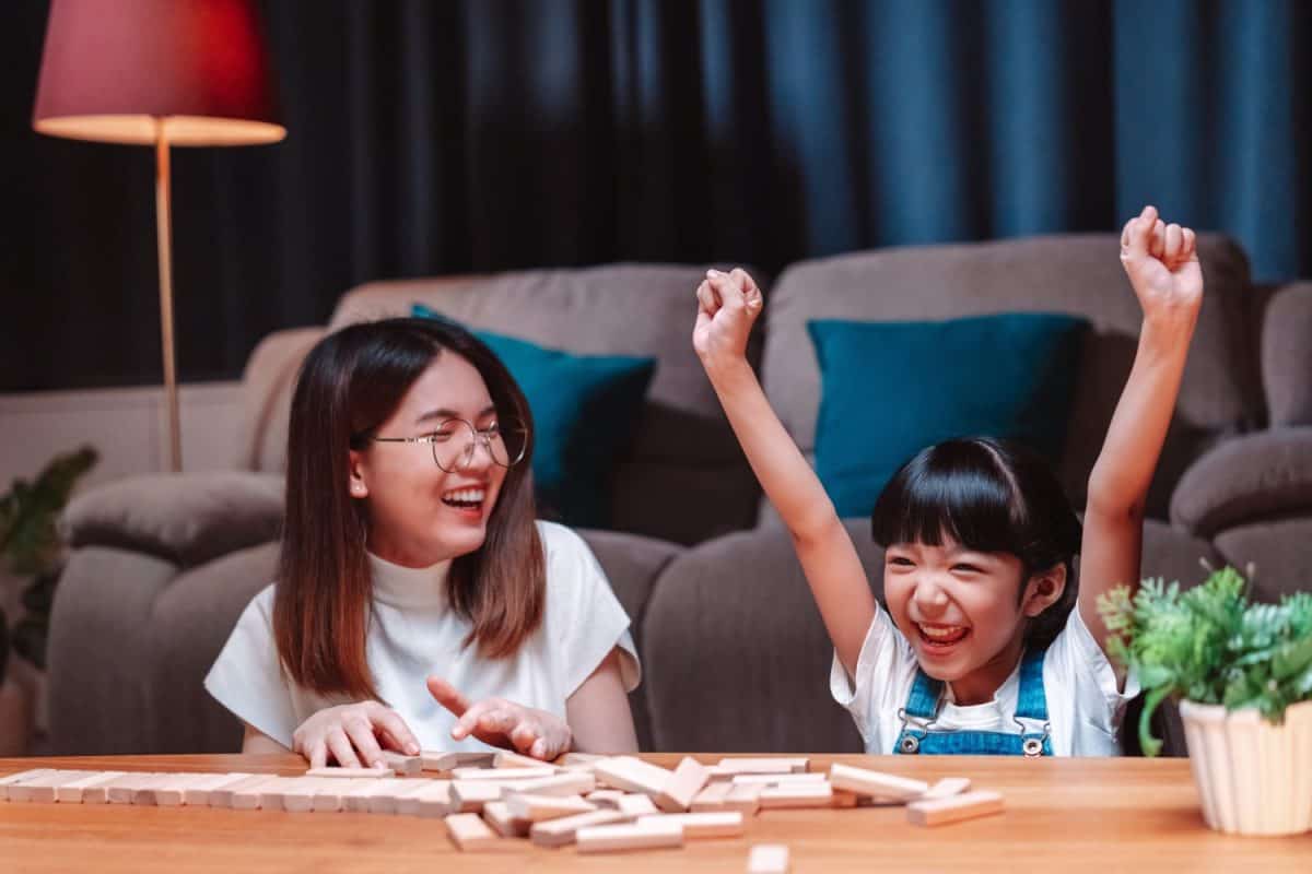 Asian happy family of little daughter play toy wooden block in home. Mother or mom young woman loving parent having fun play stack tower wood block game at night in living room. Hobbie leisure concept